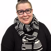 Nainen jolla on kaulahuivi kaulassa istuu ja katsoo kameraan ja hymyilee.